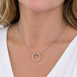 A close-up of a woman wearing a delicate silver chain necklace with a small open circular pendant adorned with tiny clear stones, styled with a white V-neck top.