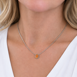 A close-up of a woman wearing a delicate silver chain necklace with a small circular pendant featuring a textured orange center and a gold-toned outer ring.