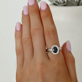 A close-up of a hand with light pink manicured nails wearing a silver ring featuring a dark oval center stone and a halo of small clear stones.