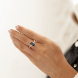 Hand wearing a ring with a blue gemstone on a neutral background