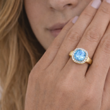 Close-up of a hand wearing a gold halo ring with a blue gemstone.