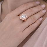 Close-up of a hand wearing a rose gold ring with a large diamond, against a soft pink background.