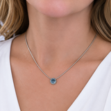 A close-up of a woman wearing a delicate silver chain necklace with a small round bezel-set light blue gemstone pendant, paired with a white V-neck top.