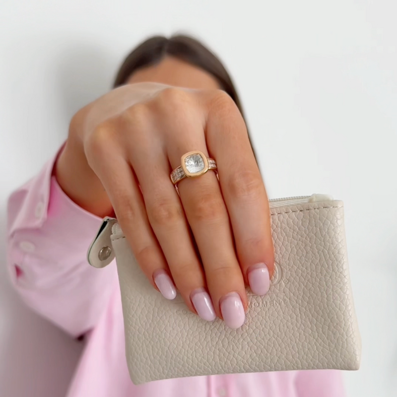 Hand holding a small beige pouch with a gold ring on a plain background