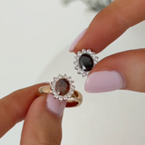 Two oval halo rings held between fingers against a soft light background. One ring has a dark brown oval center stone with clear surrounding stones and a gold band.